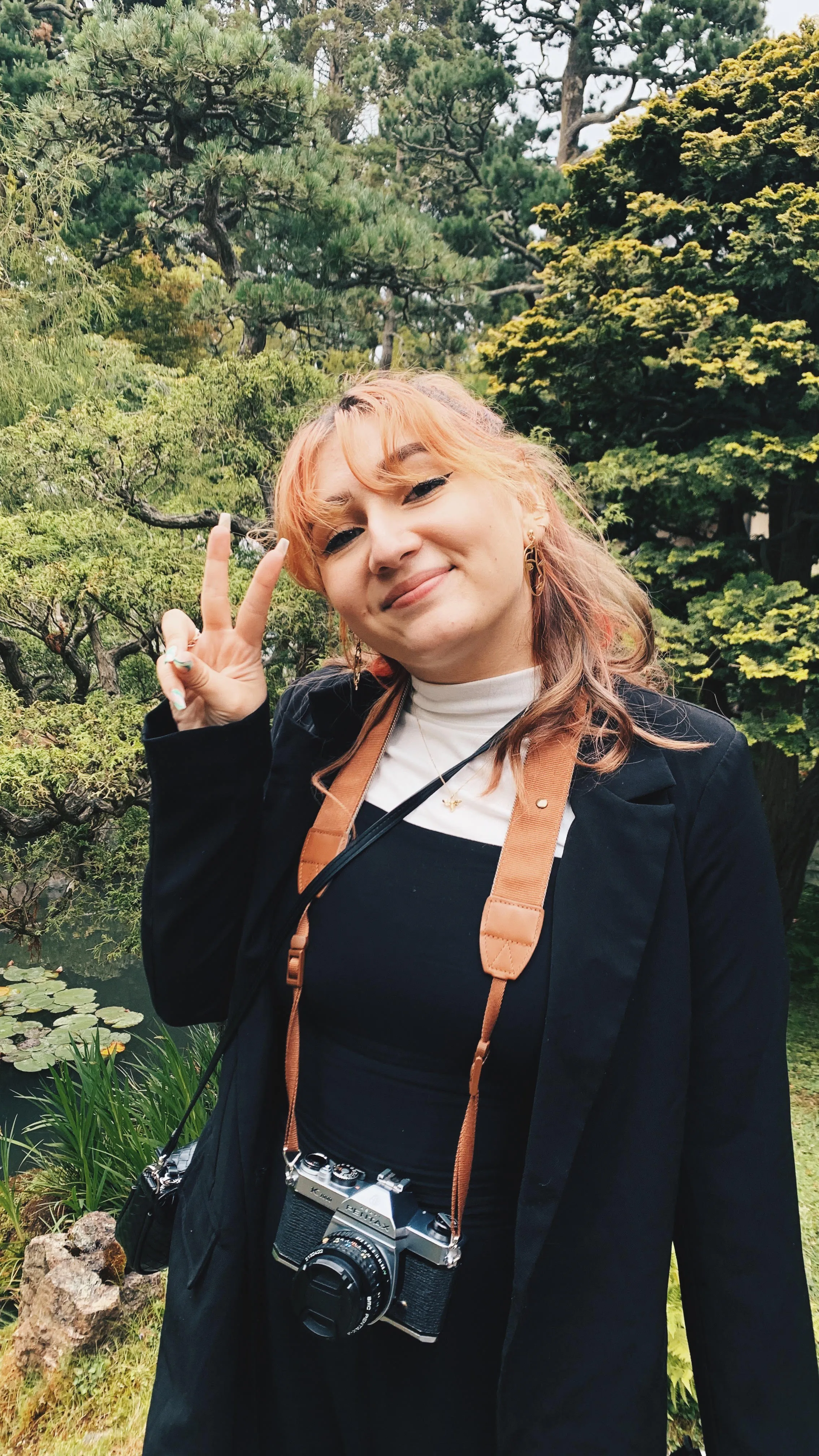 Alyssa giving a peace sign in a garden, a vintage film camera hanging from a tan strap around her neck.