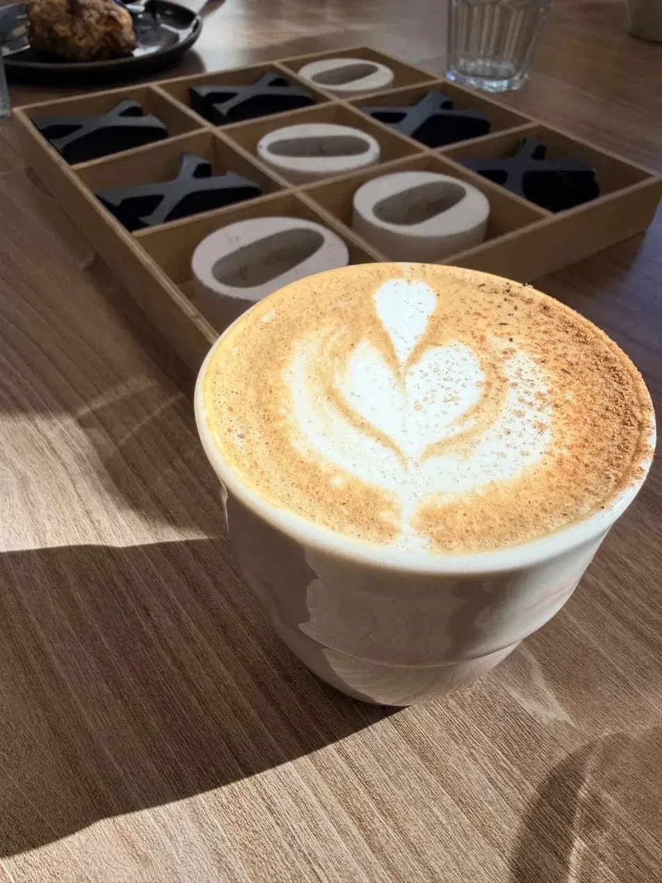 A coffee with latte art in a white cup on a sunny wooden table, next to a tic-tac-toe board.