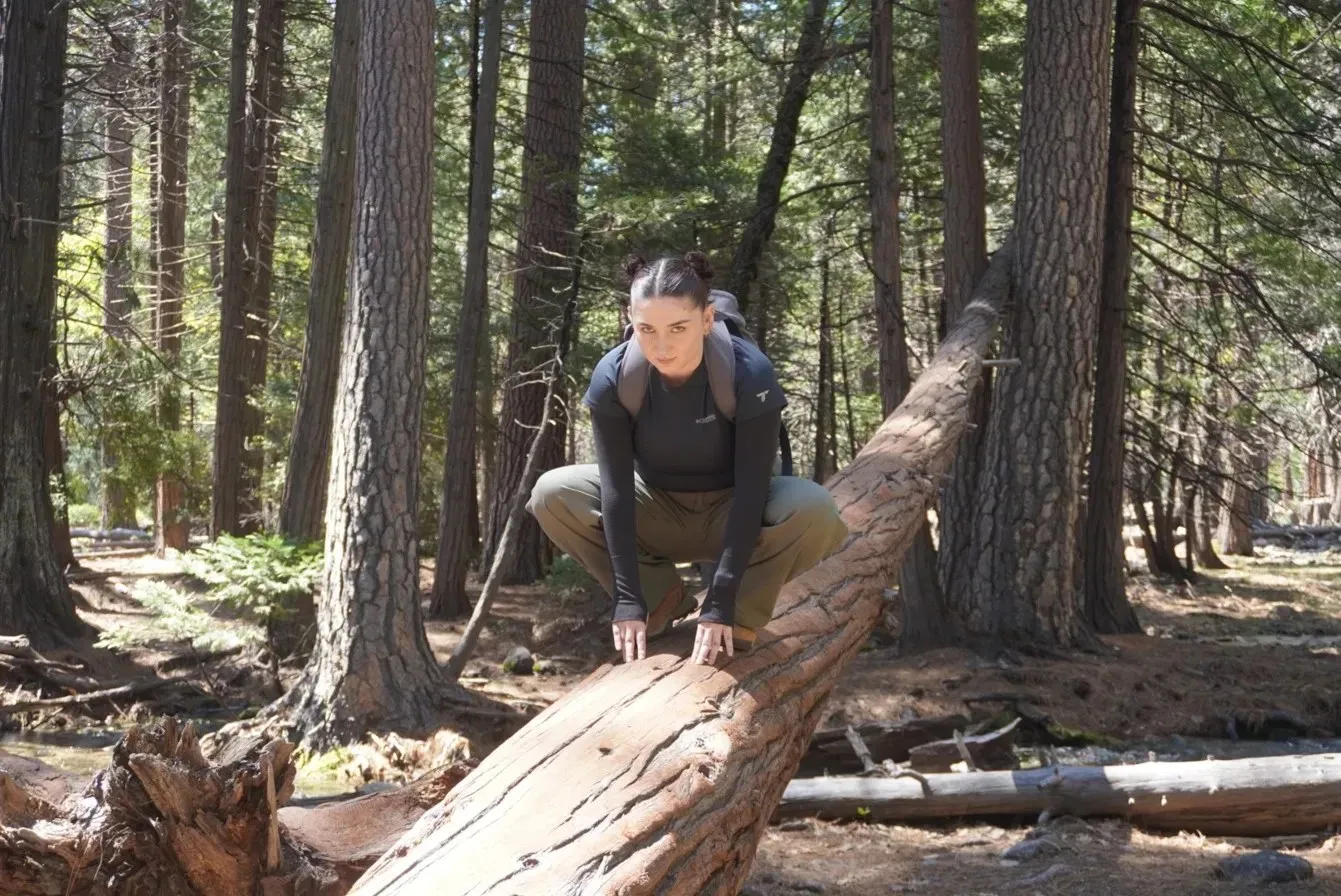 Alyssa crouched on a fallen tree trunk in a pine forest, wearing a backpack and looking at the camera.