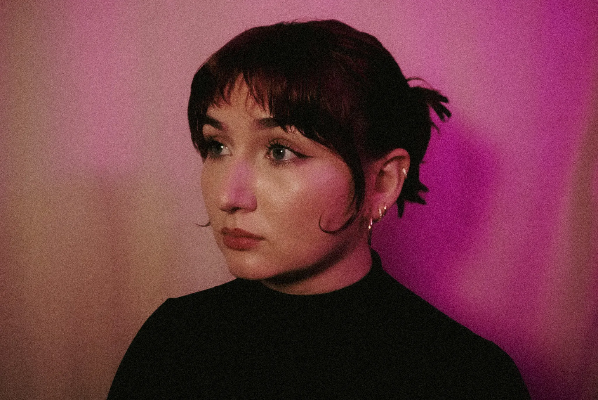 Alyssa in a portrait lit in pink and magenta, looking off to the side, in a black turtleneck against a rose wall.