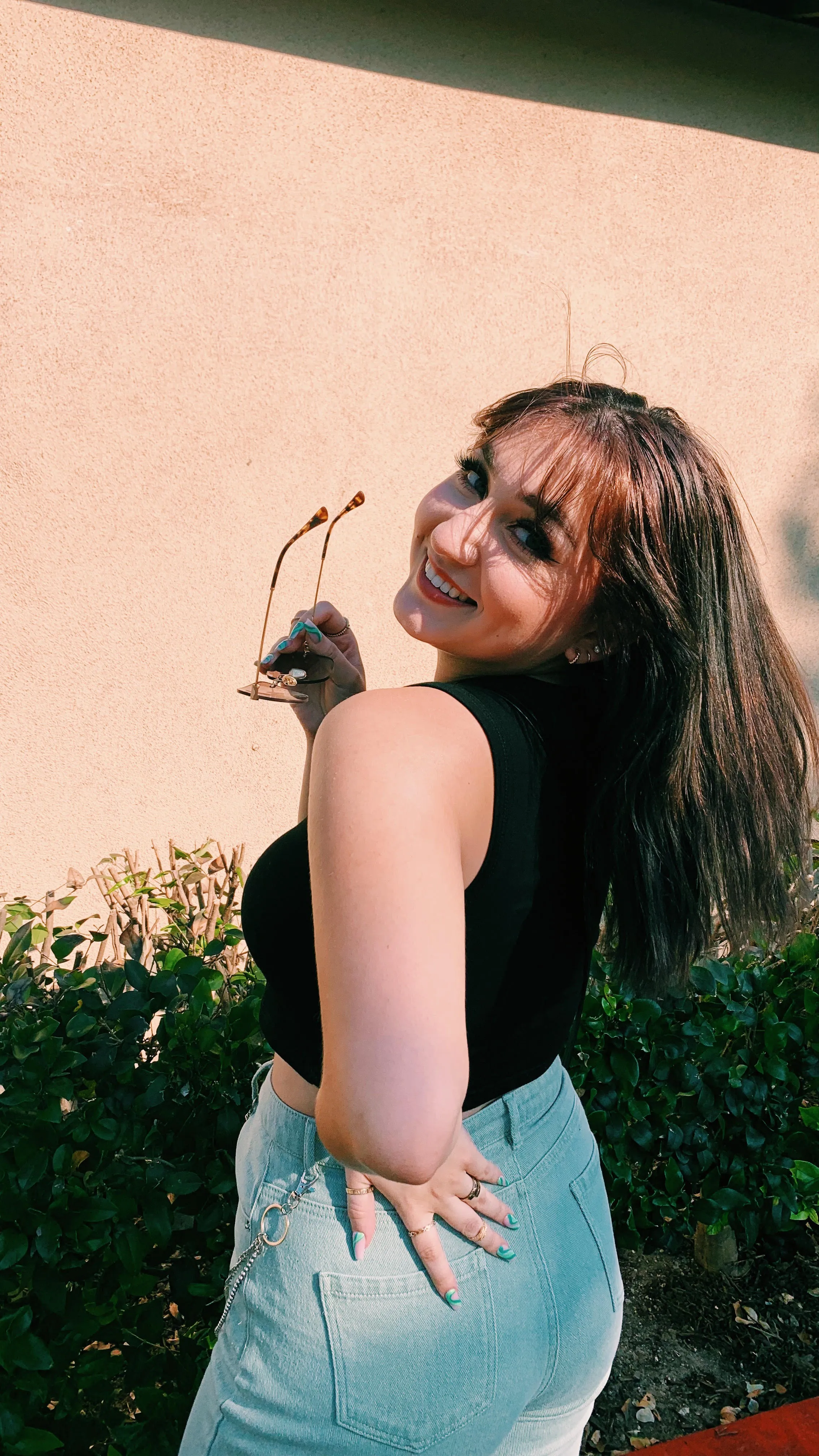 Alyssa looking back over her shoulder and smiling, holding sunglasses, in warm light against a peach wall.